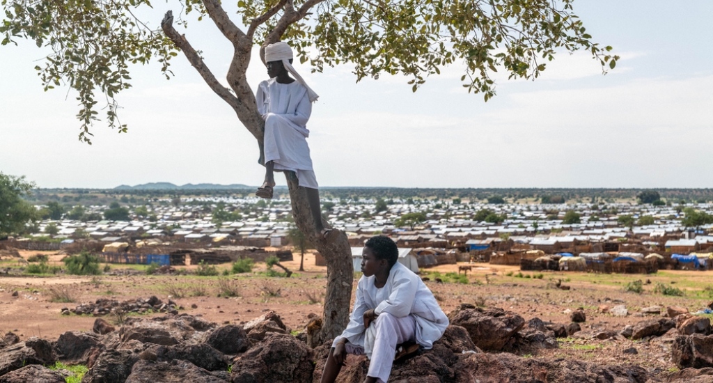 Twee Soedanese jongens rusten op een heuvel met uitzicht op het uitgestrekte Aboutengue-vluchtelingenkamp, dat onderdak biedt aan tienduizenden mensen die aan etnisch geweld in West-Darfur, Soedan, zijn ontsnapt.  Ouaddaï-provincie, Tsjaad, juli 2025.